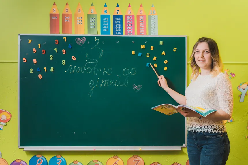 A female teacher pointing to a chalkboard with numbers in a colorful classroom.