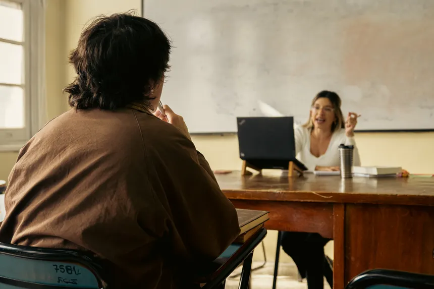 Student and teacher engage in discussion in a classroom setting with a laptop and whiteboard visible.